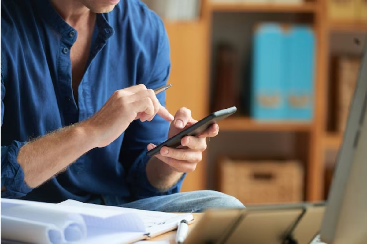 Smartphone: Aplicativos para ganhar dinheiro Homem usando Aplicativos para ganhar dinheiro em smartphone dentro de sua casa e analisando dados em papéis em sua frente