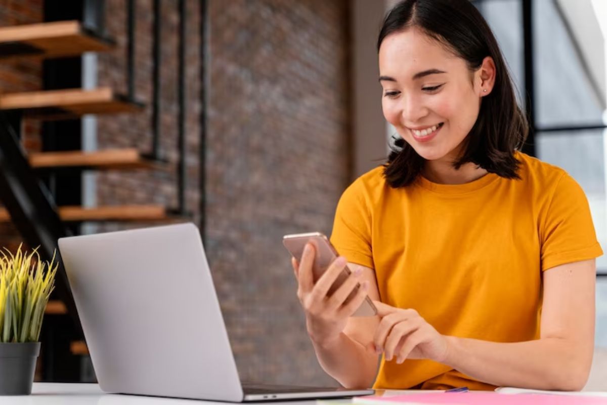 Mulher sentada diante de notebook usando Aplicativos para ganhar dinheiro no celular sorridente em interior de casa