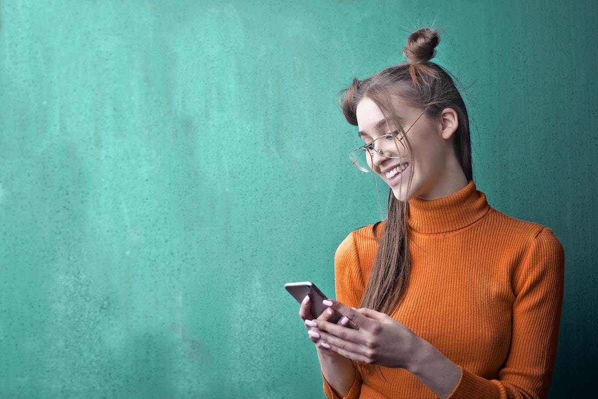 Mulher usando Apps que pagam por cadastro em celular diante de parede verde e sorridente