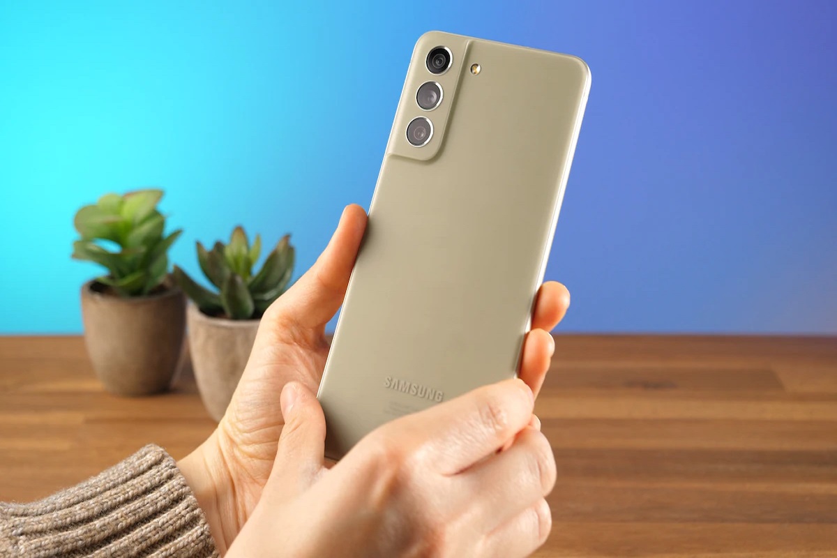 Análise: Galaxy S21 FE Galaxy S21 FE com pessoa segurando como se analisasse o aparelho com as duas mãos com mesa de madeira com plantas de interior ao fundo em desfoco na imagem