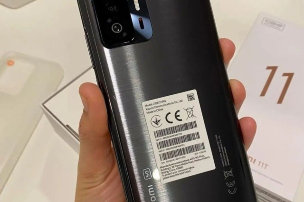 Pessoa segurando aparelho Xiaomi 11T preto com as costas para cima como se mostrasse o mesmo durante review em vídeo e com caixa e acessórios ao fundo em desfoco