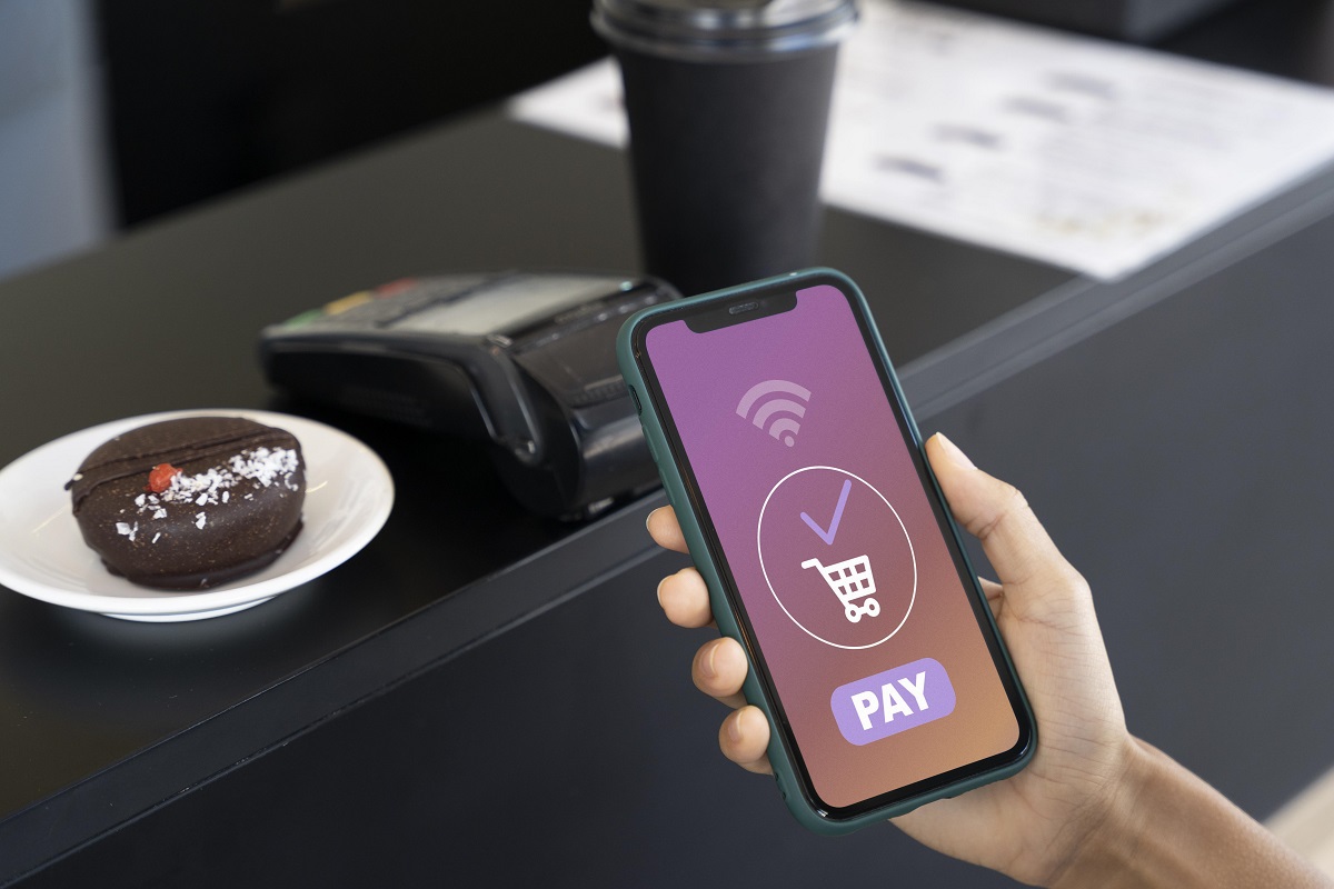 Celular: Aplicativos de bancos Pessoa segurando celular usando um dos Aplicativos de bancos para pagamento online diante de doce sobre pires e ao lado de pinpad