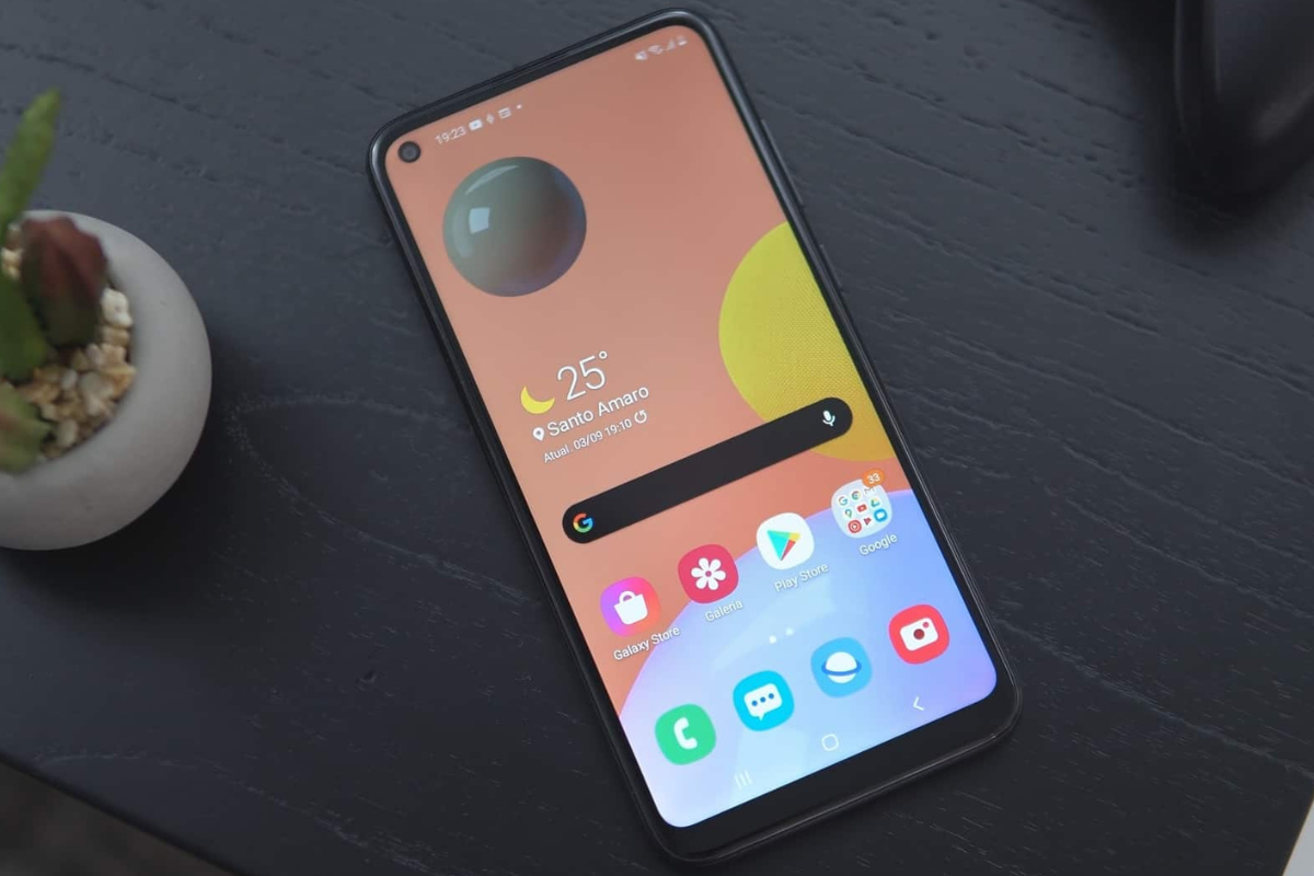 Tela: Galaxy A11 Galaxy A11 sobre superfície escura e ao lado de planta de interiores enquanto aparelho mostra sua tela inicial