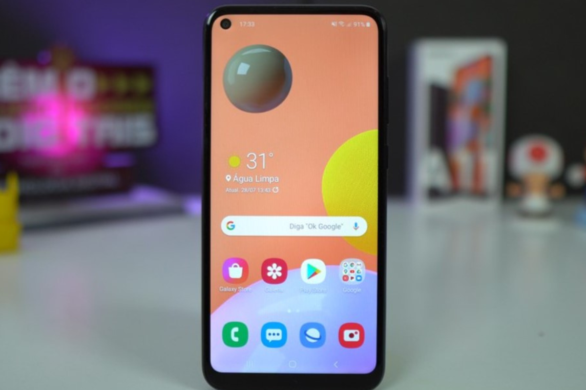 Review: Galaxy A11 Galaxy A11 em suporte sobre mesa com sua caixa e outros objetos ao fundo em review
