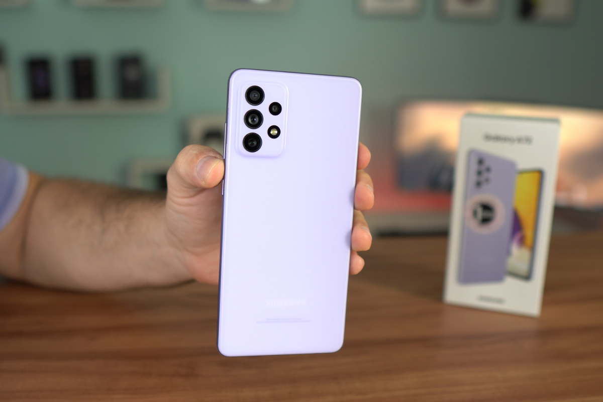Câmeras: Galaxy A72 Usuário segurando Galaxy A72 diante de mesa de madeira com caixa do produto sobre a mesma mostrando as costas do aparelho e suas câmeras