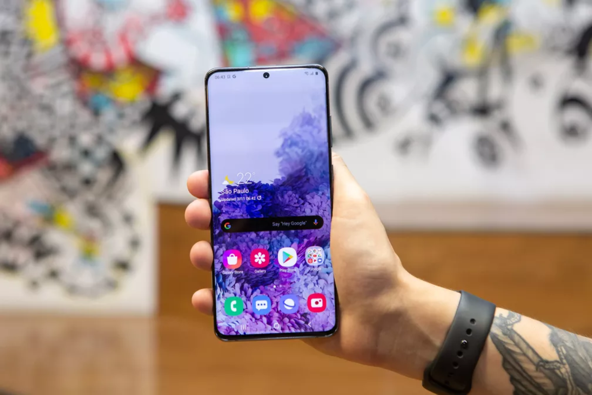 Usuário segurando celular modelo Galaxy S20 Ultra com mão direita enquanto mostra sua tela em menu inicial desbloqueado para câmera