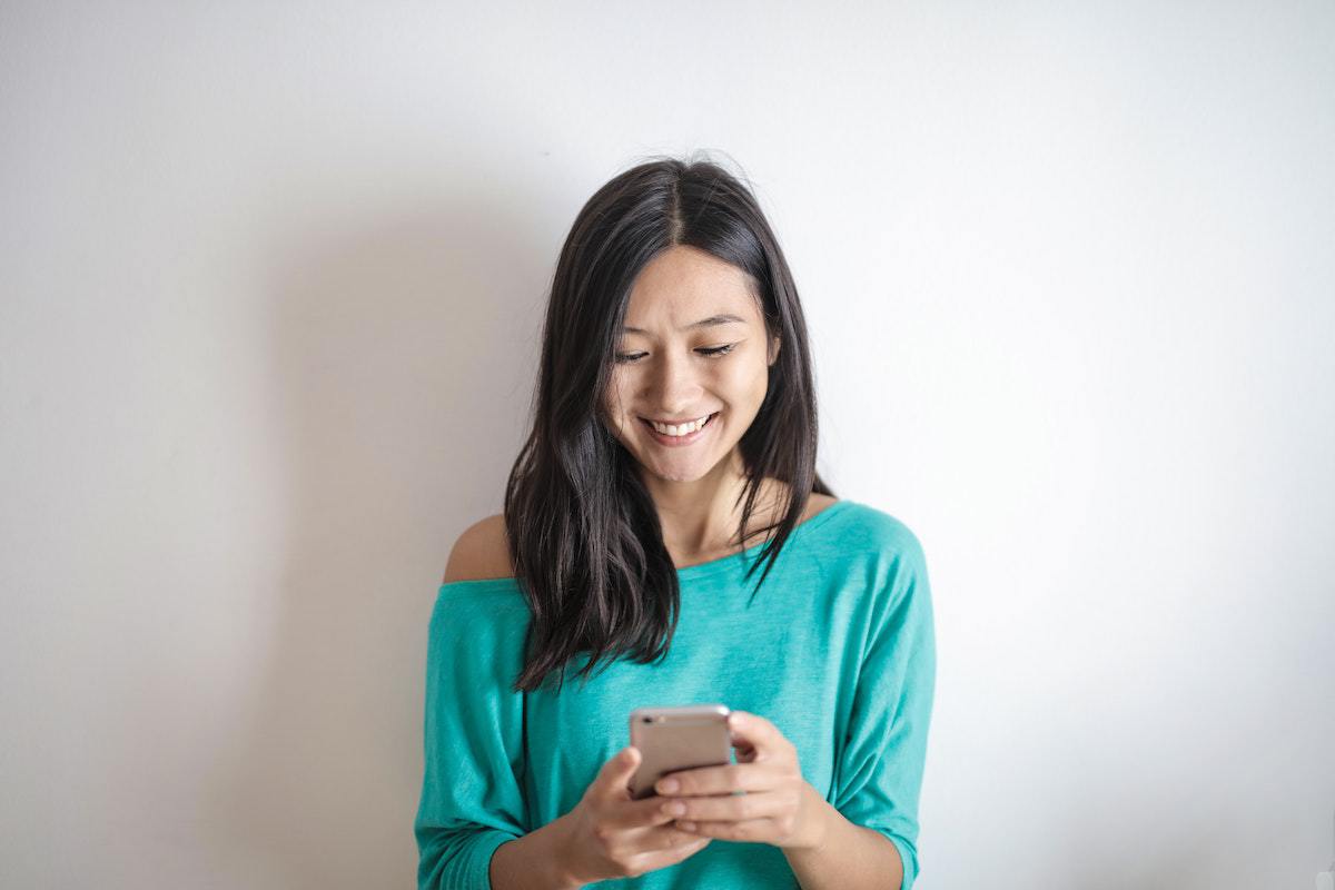 Celular: Smartphones Mulher usando celular sorridente enquanto aparentemente digita algo escorada em parede