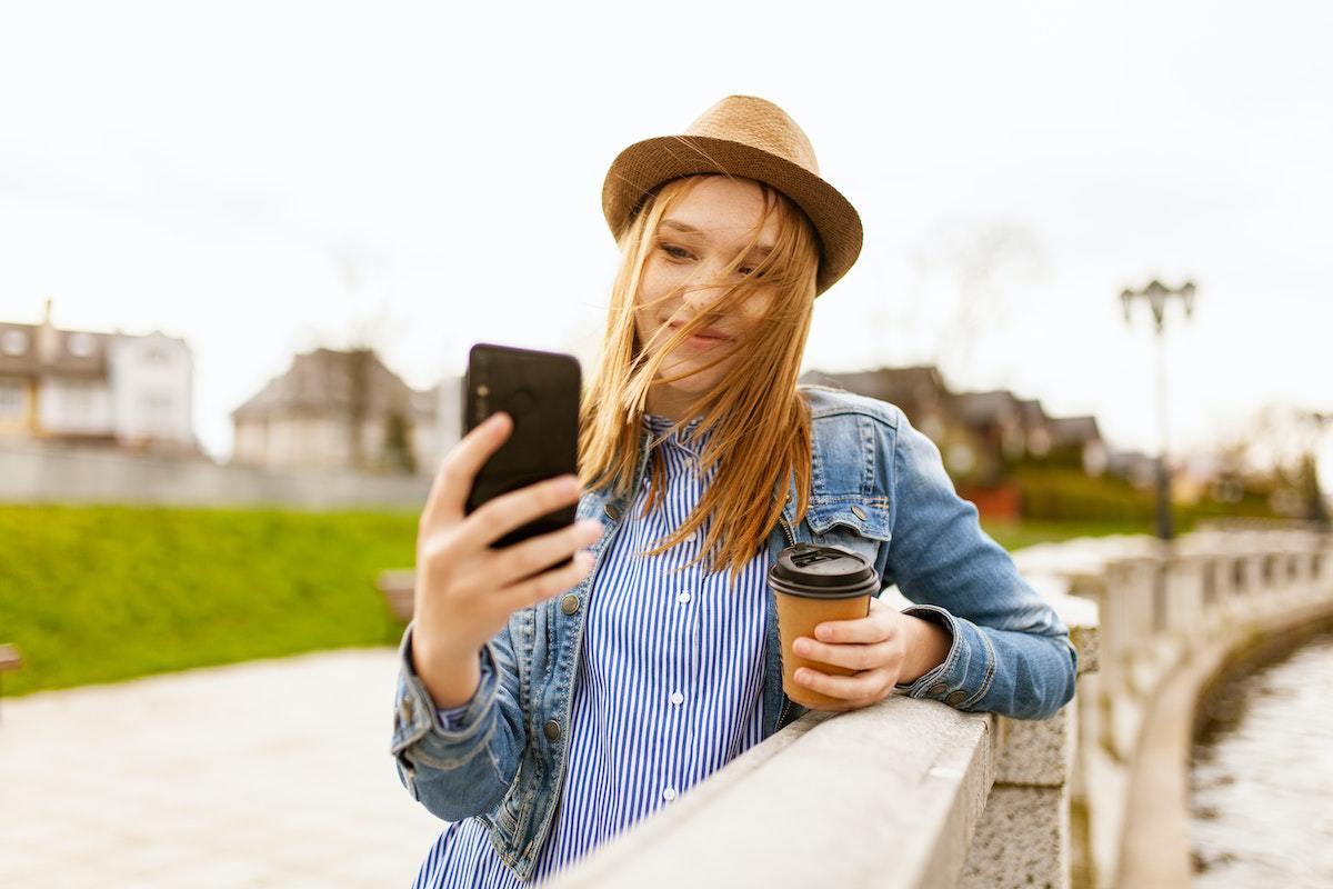 Selfie: Smartphones Mulher usando chapéu e camisa social azul encostada em pilares de local que parece ponte tirando uma foto tipo selfie com seu celular com um copo de café em mãos