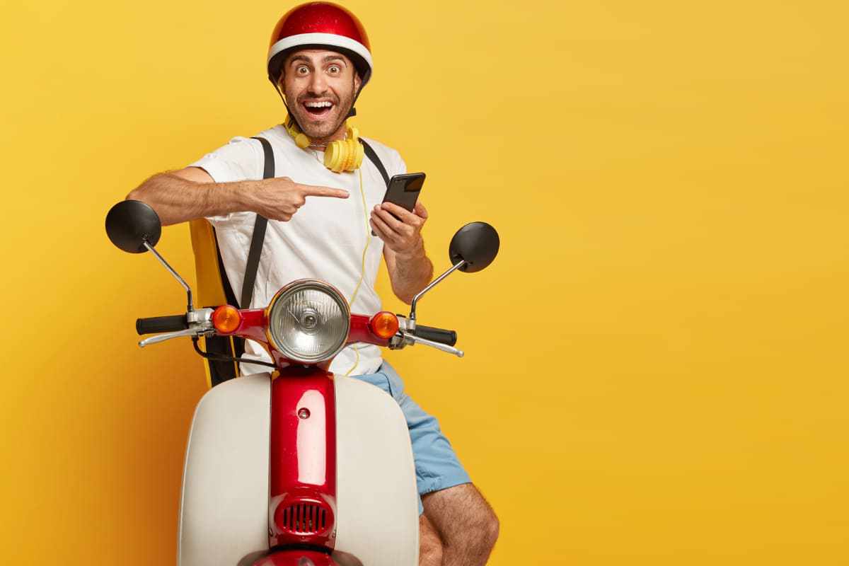 aplicativos de entrega: a imagem tem um fundo amarelo, além disso, está um homem em cima de uma motocicleta apontando para um smartphone.
